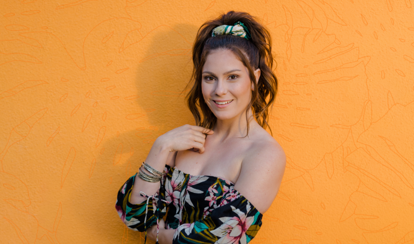 An elegant woman wearing a floral outfit, posing against an orange wall with her hair in a ponytail.