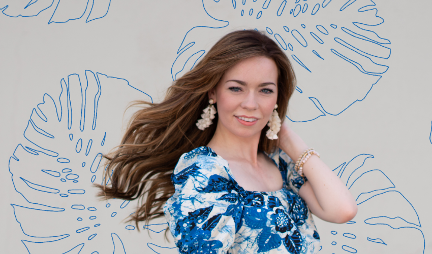 A woman in a blue floral dress, with hair extensions, posing with a blue monstera leaf behind her.
